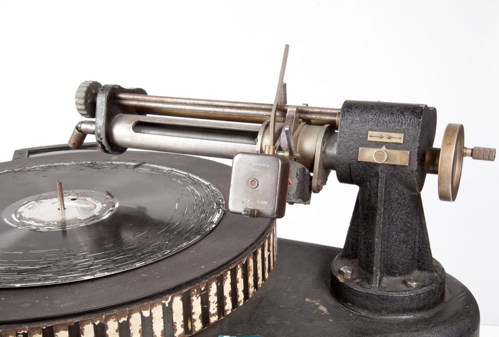 Lähikuvassa Yleisradiolla käytössä ollut äänilevyleikkuri. Äänilevyleikkuri mahdollisti ensimmäisenä radio-ohjelman tai sen osan tallentamisen. Musta metallinen laite, valkoisella taustalla.