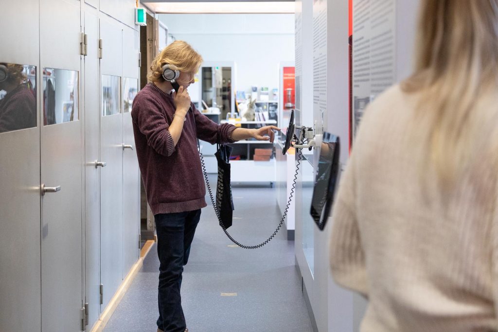 Museovieras kuuntelee kosketusnäytön sisältöjä näyttelytilassa.